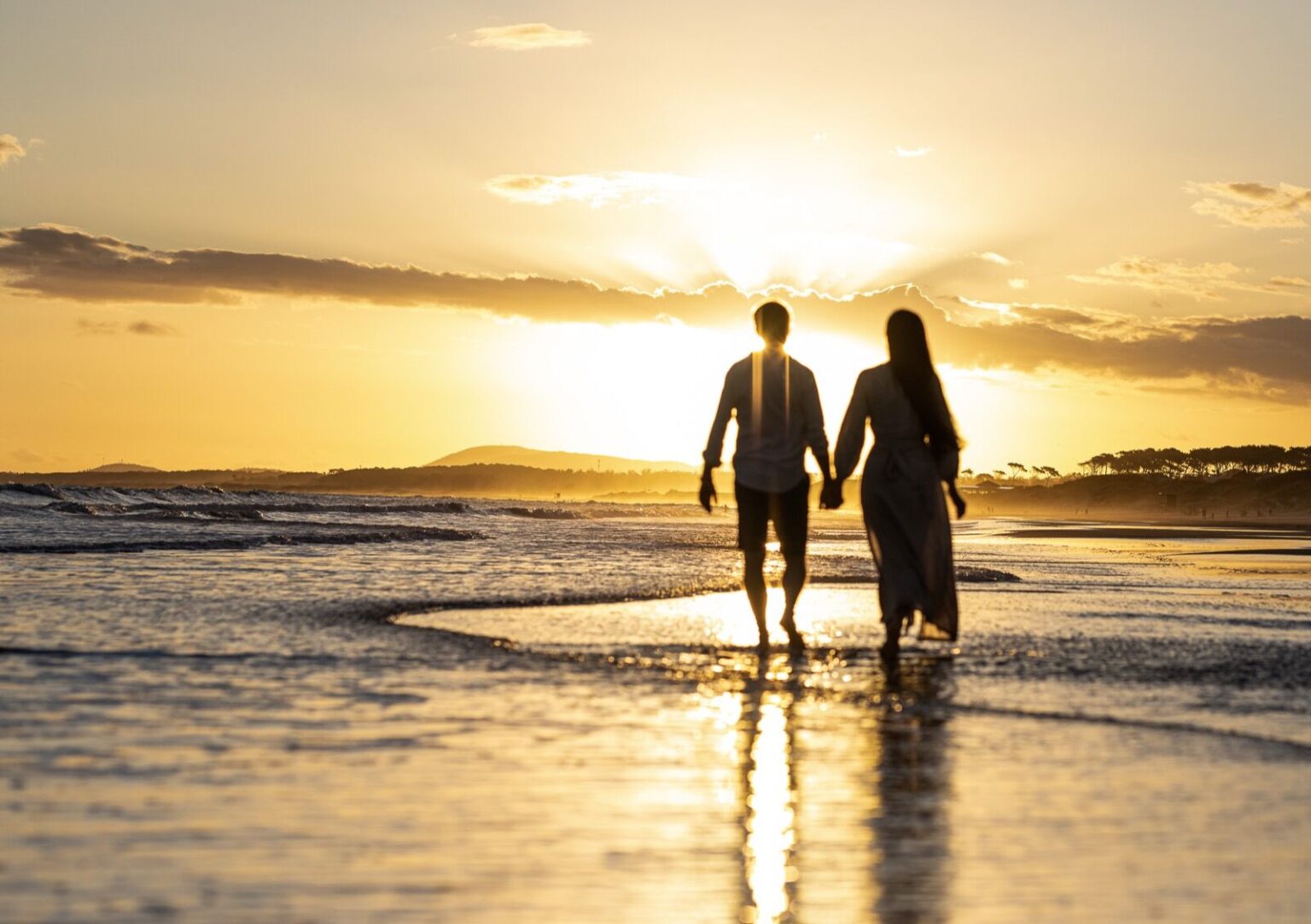 Los novios caminan de la mano por la orilla al atardecer, con el sol detrás