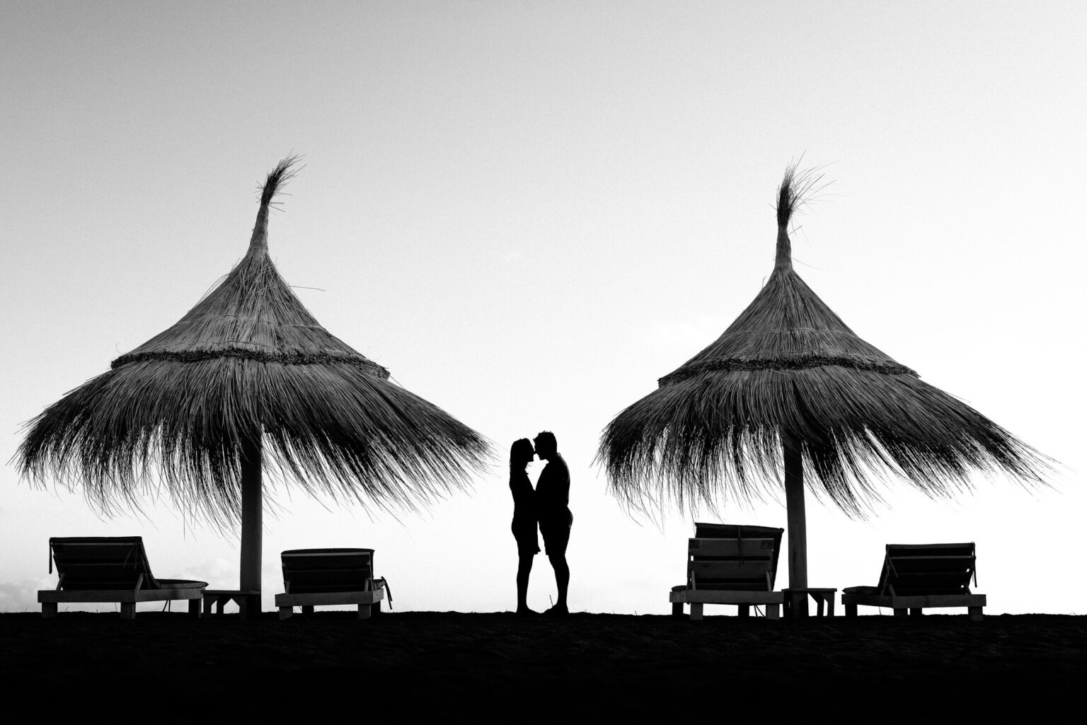 Beso entre sombrillas de paja frente al mar, en blanco y negro