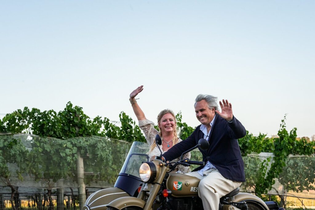 Los novios saludando desde una moto con sidecar entre viñedos